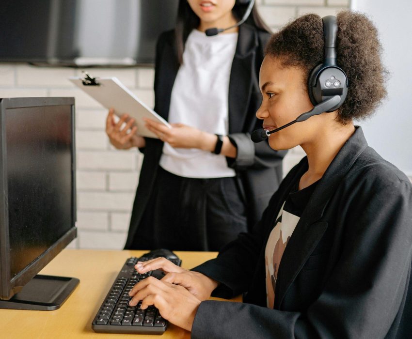 Two women in a call center wearing headsets, working at a computer.